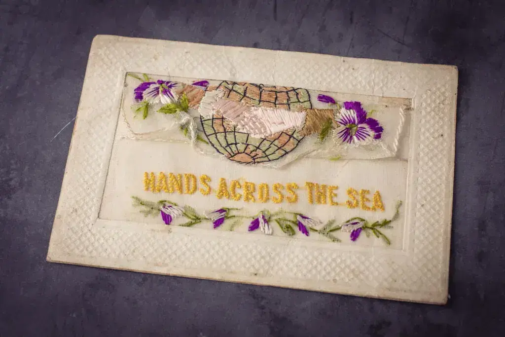 Vintage embroidery war postcard on lace of flowers and two hands shaking in front of the globe with the script hands across the sea underneath